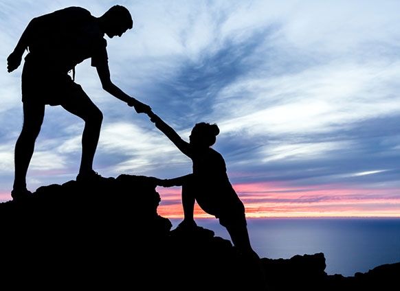 two people are helping each other up on top of a mountain with the sun setting in the background