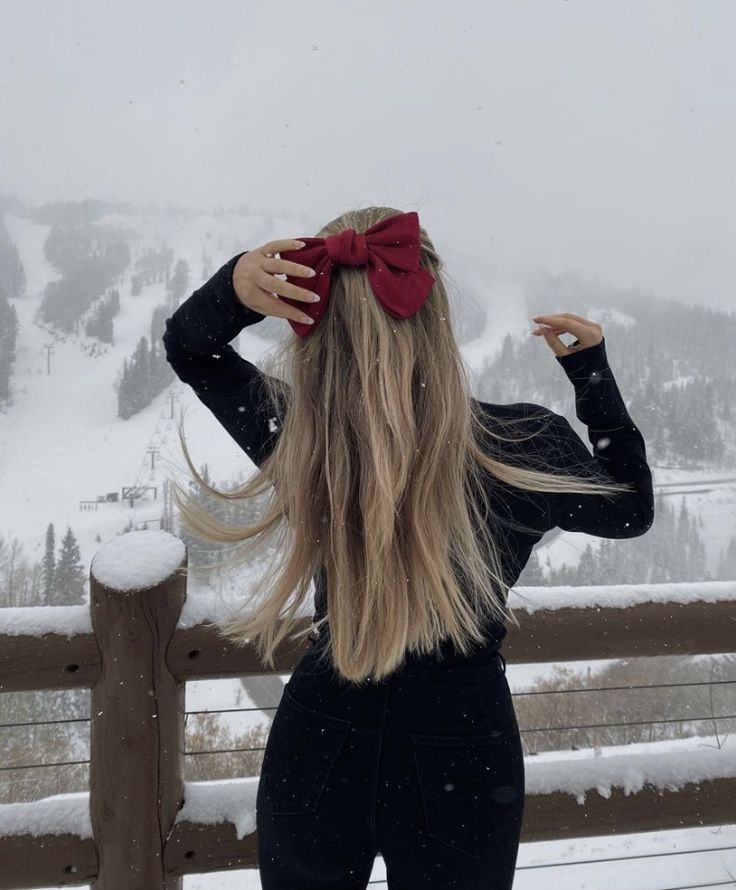 a woman with long blonde hair wearing a red bow in front of snow covered mountains