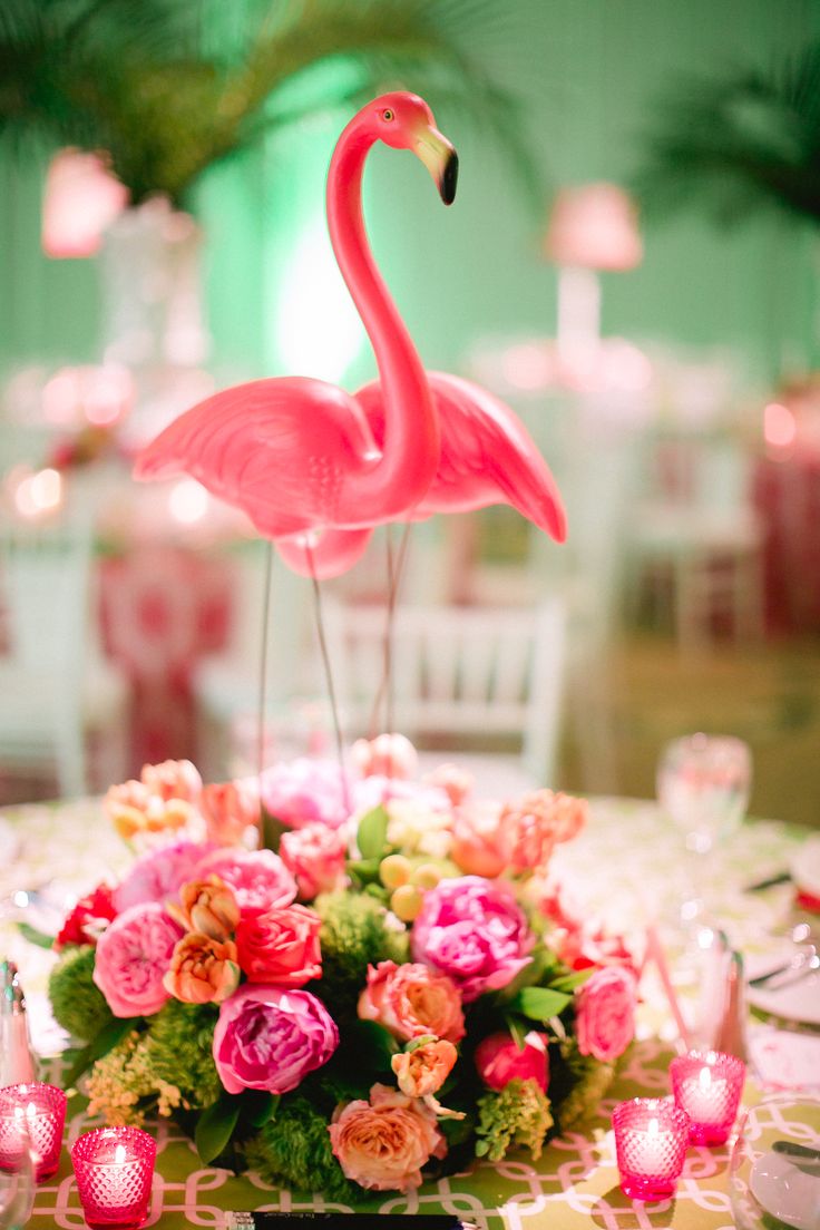 a pink flamingo sitting on top of a table next to flowers and candles in vases