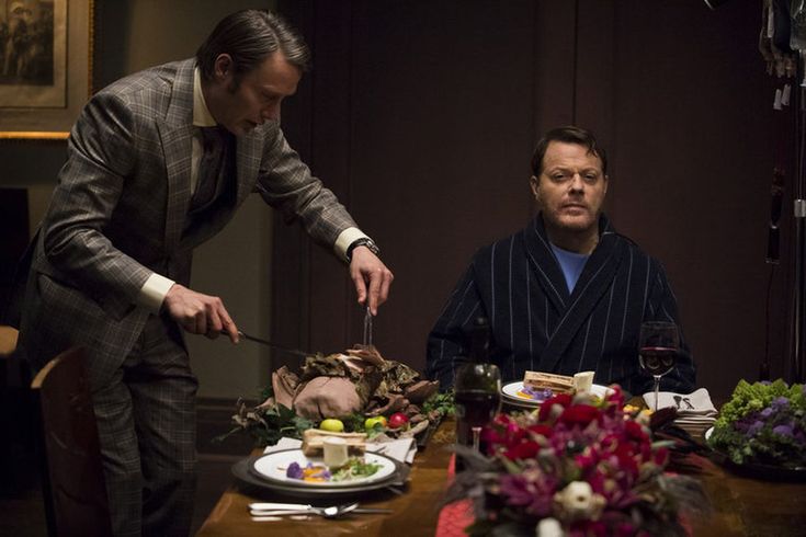 two men standing at a table with food in front of them and one man holding a knife