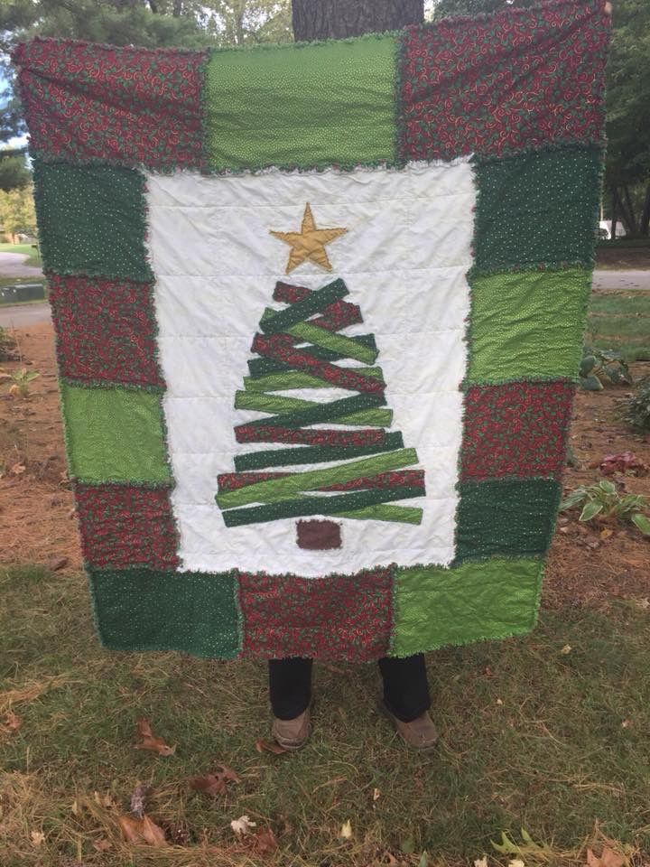 a person standing in front of a christmas tree quilted on the ground, holding up a green and white blanket