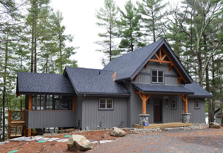 a gray house in the woods with lots of pine trees and rocks on the ground