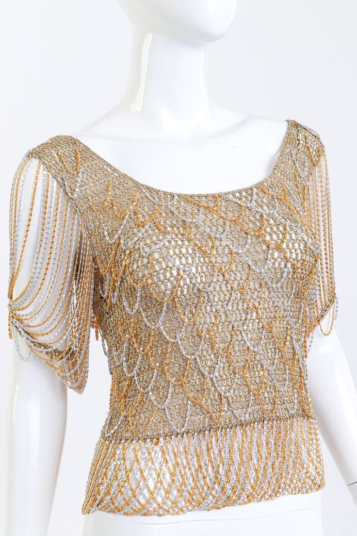 Loris Azzaro gained international fame for dressing celebrities like Tina Turner in his glimmering textiles. Items such as this gold metallic knit top featuring draped chain details have since become fashionista collectors pieces. Shimmering sheer short sleeve knit and chain top Gold, silver Circa early 1970s Lamé woven knit & fine spiral chain drape detail Scoop neckline Chandelier chain sleeves Criss-cross draped hem Pullover style No fabric contents - silk/wool mix, metallic fibers Loris Azzaro label, made in France Very good vintage condition, stained label Estimated Size: S Measurements ~has up to 4* of stretch Bust: 28"-32"Waist: 25"-29" Length (Centre Front to Waistband): 10.5"Waistband (Chain Drop): 4"Length (Centre Back): 16"Shoulder-to-Shoulder: 16"Sleeve Length: 8" Sleeve Openin Gold Short Sleeve Top With Sequins, Gold Short Sleeve Tops With Sequins, Gold Sequined Short Sleeve Top, Gold Sequin Short Sleeve Top, Glamorous Gold Short Sleeve Top, Chic Embellished Gold Tops, Formal Embellished Gold Tops, Glamorous Embellished Gold Tops, Glamorous Gold Embellished Top