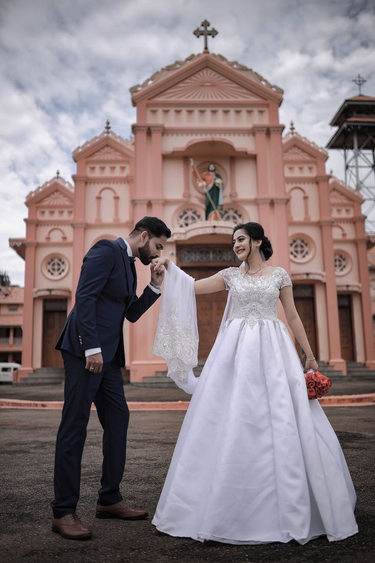 a bride and groom standing in front of a pink church with their arms around each other