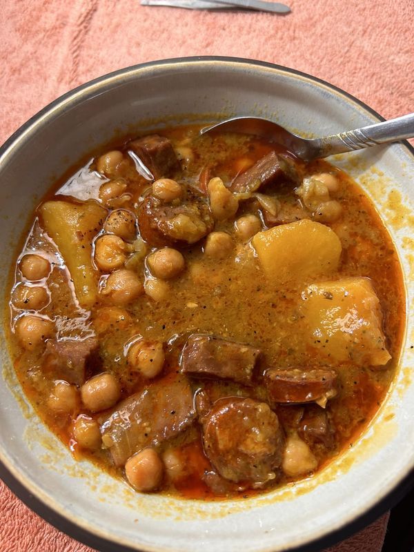 a bowl of stew with meat, beans and potatoes on a table next to a spoon