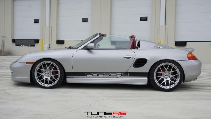 a silver porsche sports car parked in front of a garage with its doors open and the roof down