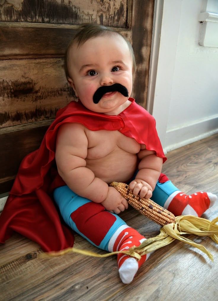 a baby dressed as a superhero sitting on the floor with a fake corn cob in his mouth