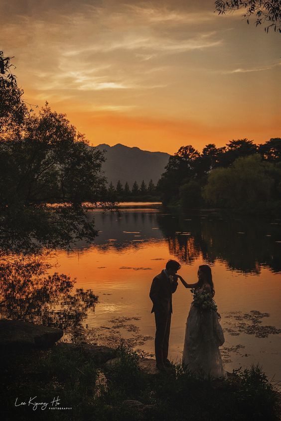 a bride and groom standing in front of a lake at sunset