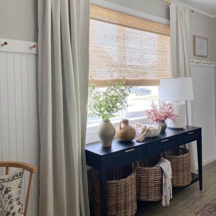 a table with baskets and vases on it in front of a window covered by curtains