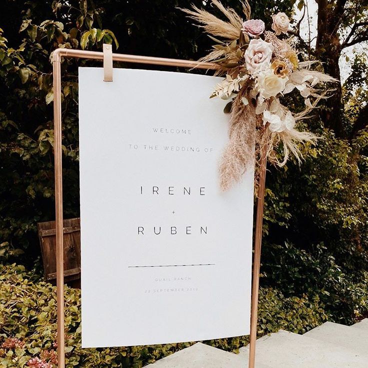 a wedding sign with flowers on it in front of some bushes and trees at an outdoor venue