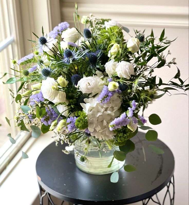 a vase filled with white and blue flowers on top of a table next to a window