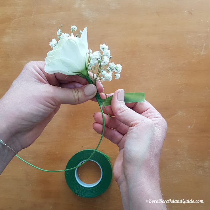 two hands are holding a white flower with green tape on the table next to it