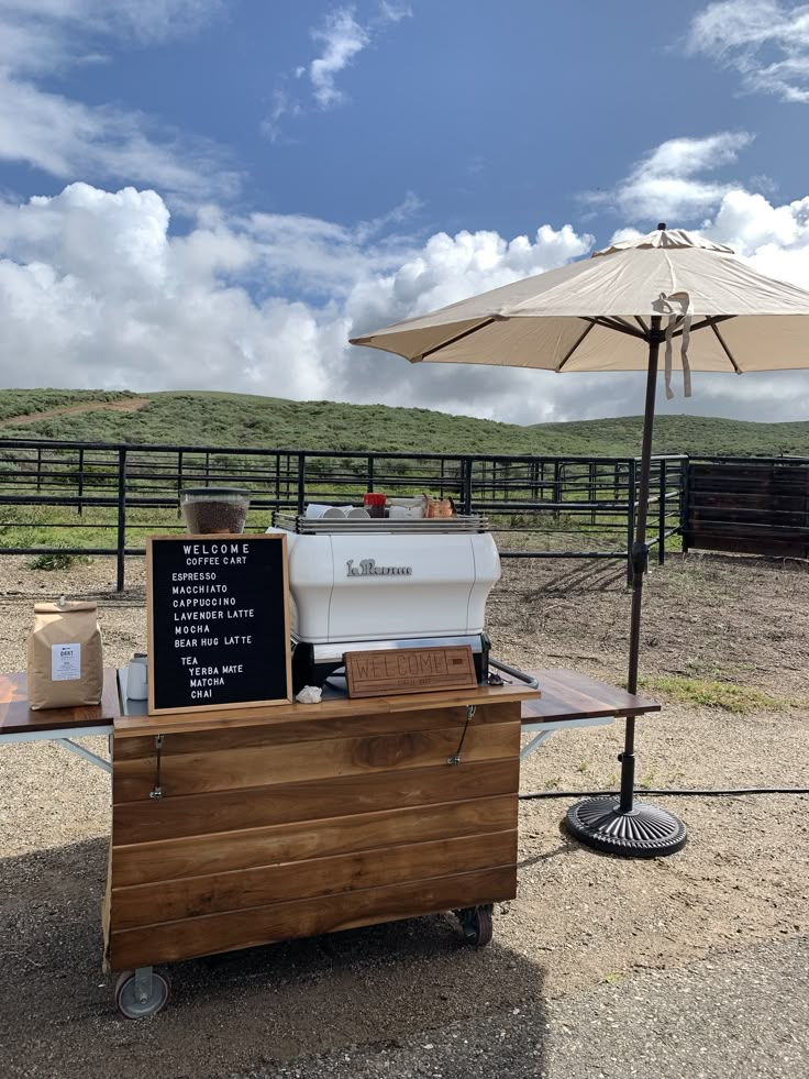 an outdoor food cart is set up in front of a fenced area with a large umbrella