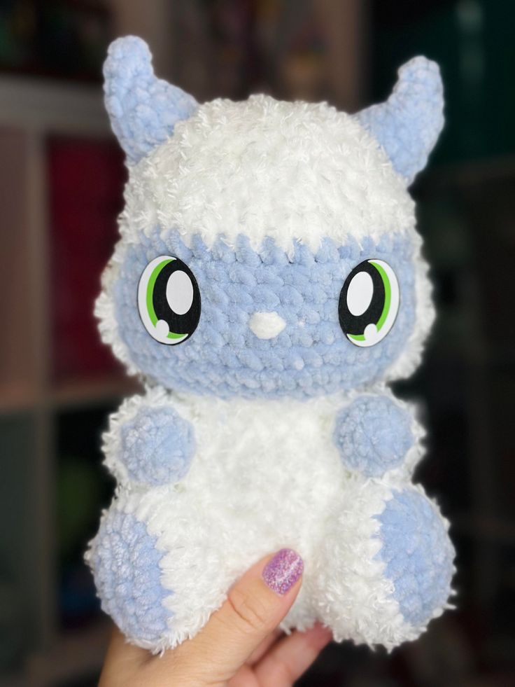 a hand holding a small white and blue stuffed animal with big eyes on it's face