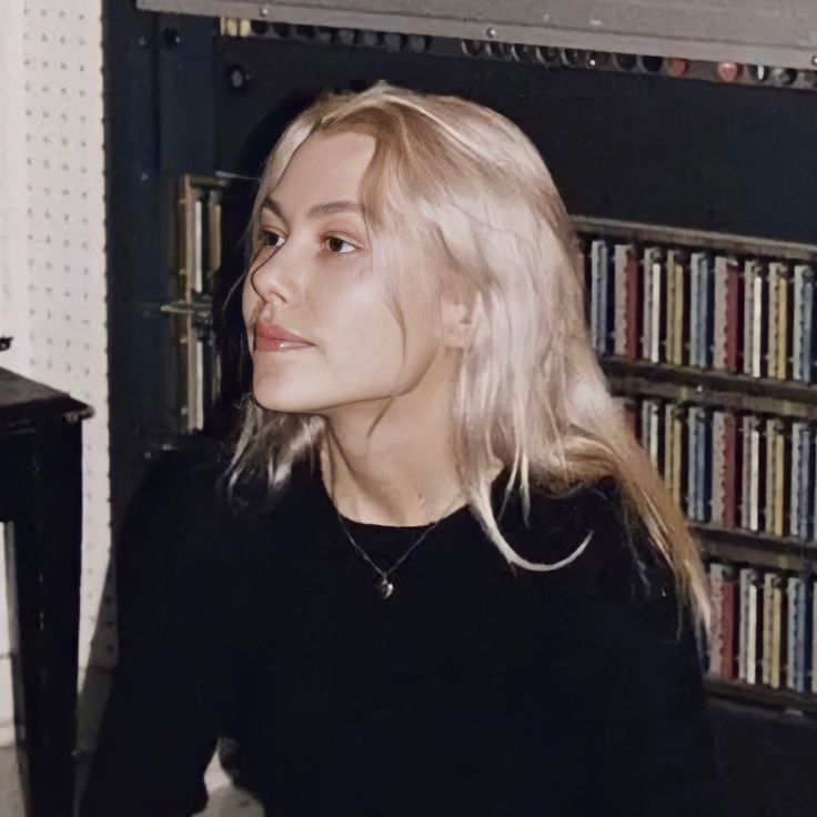 a woman with blonde hair sitting in front of a book shelf and looking off into the distance