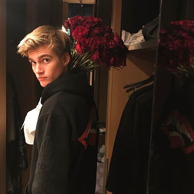 a young man standing in front of a mirror with flowers on his head and wearing a black hoodie