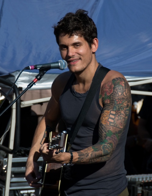 a man with tattoos on his arm holding a guitar and standing in front of a microphone