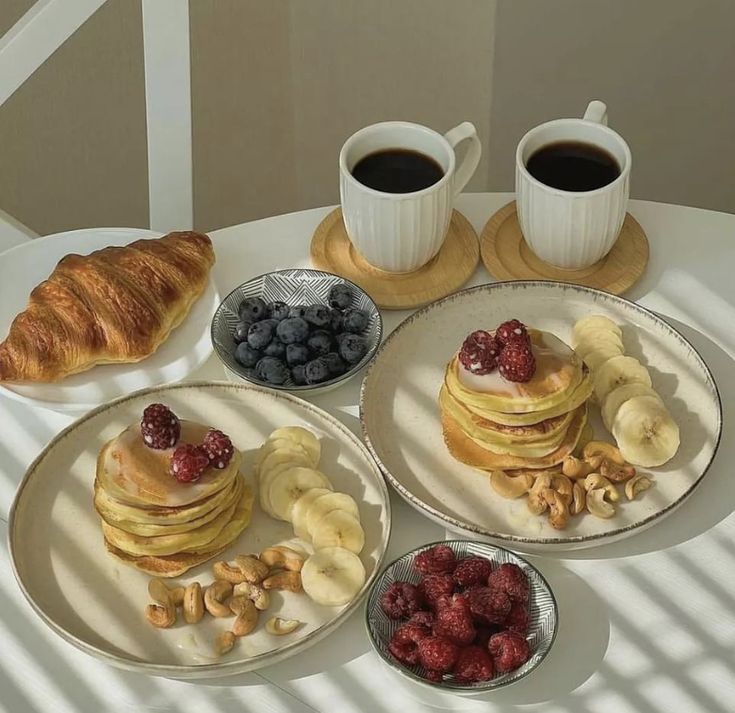 two plates with pancakes, bananas and berries on them sitting next to cups of coffee