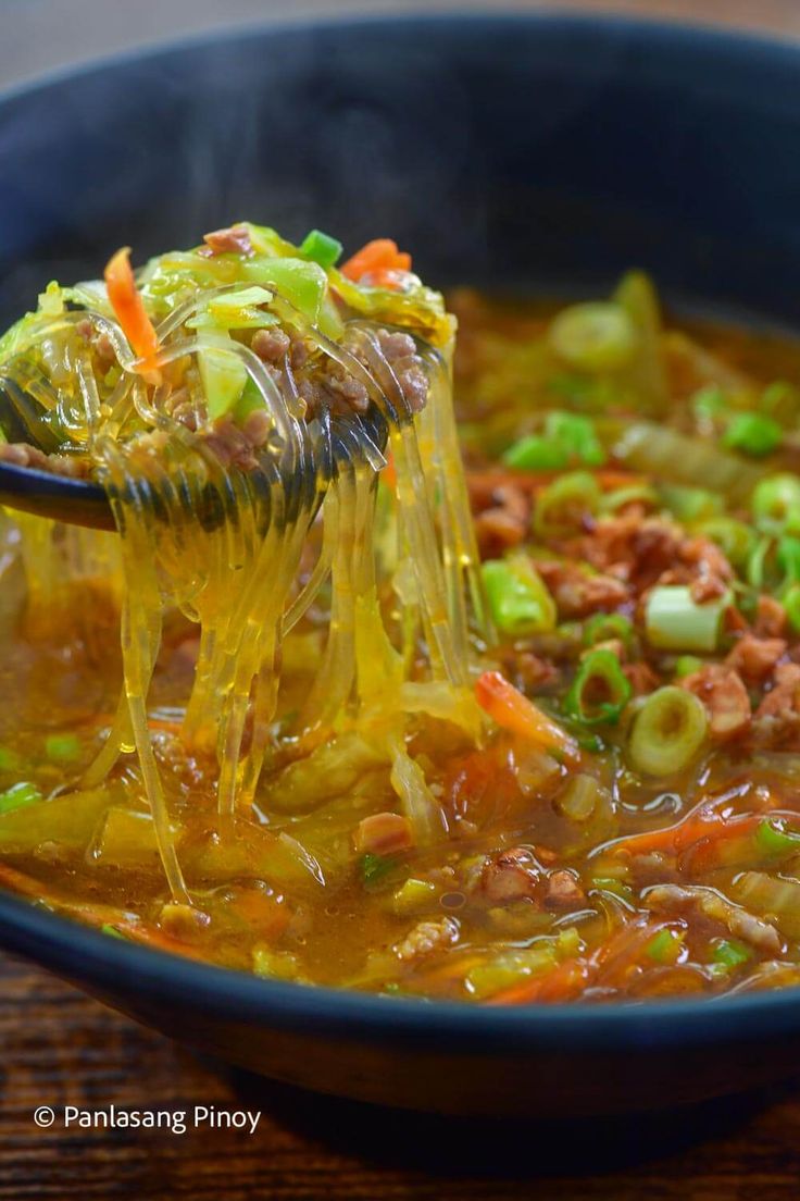 a spoon full of soup with vegetables being held up by someone's hand over the bowl