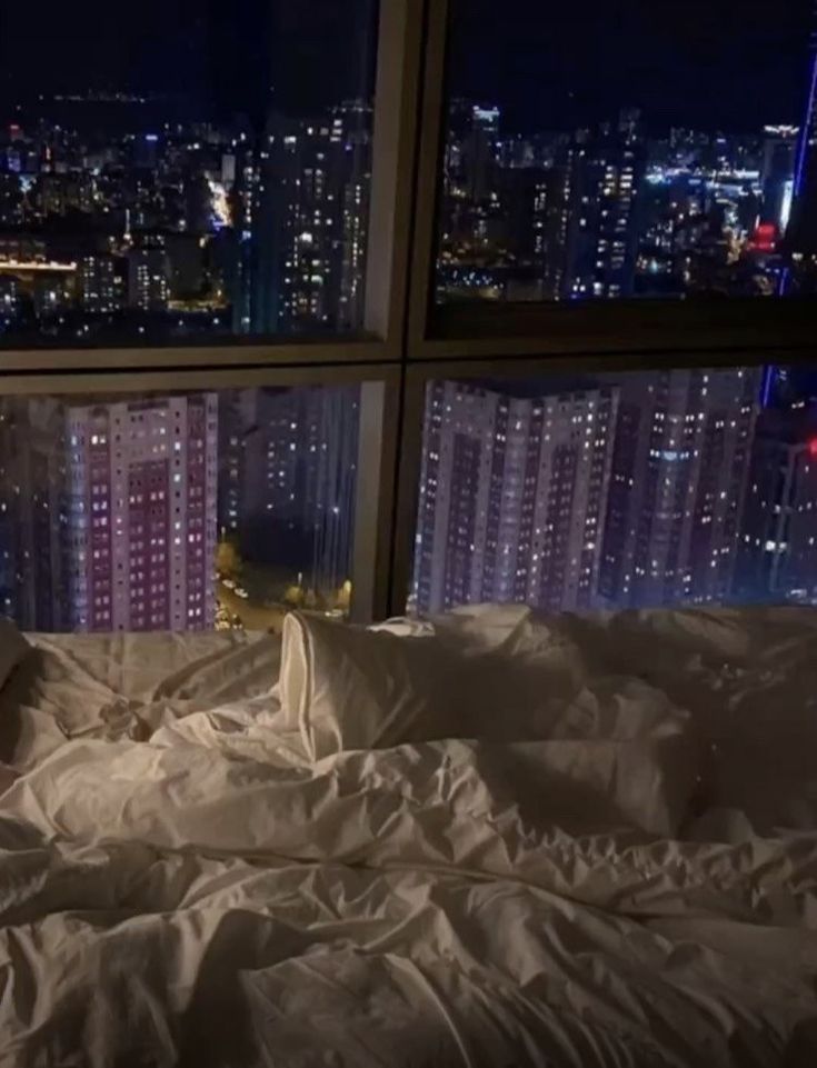 an unmade bed in front of a large window with city lights lit up at night