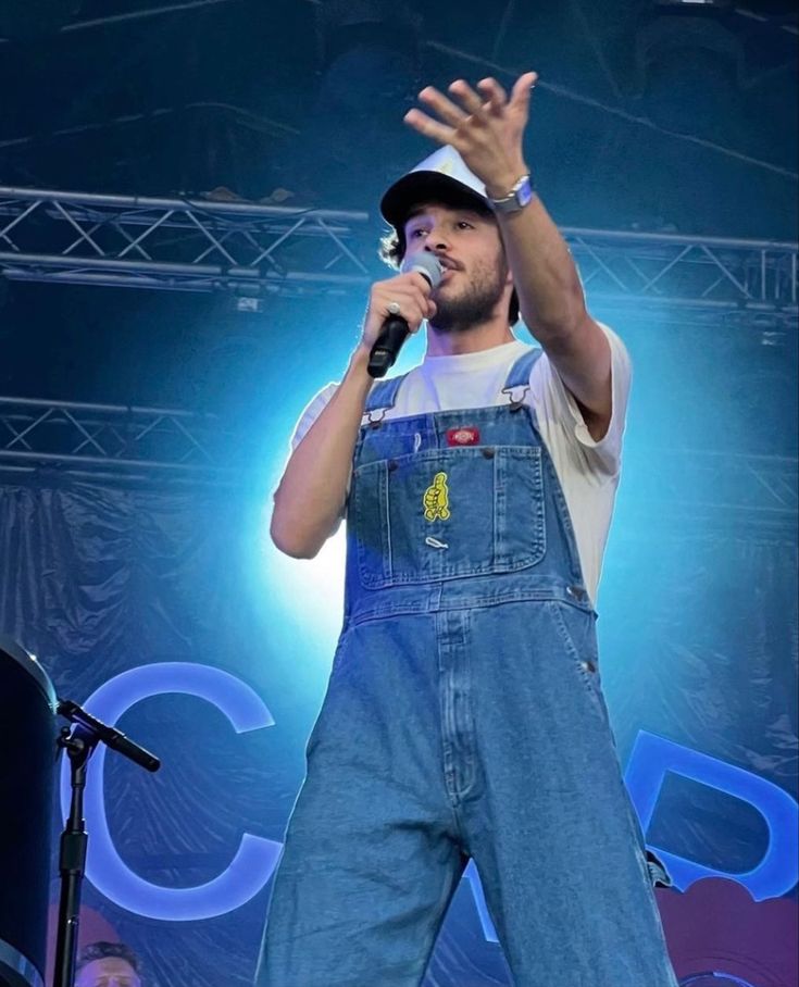 a man in overalls singing on stage with his hands up to the sky and holding a microphone