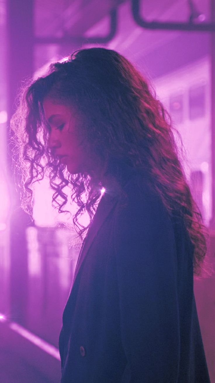 a woman with long curly hair standing in front of a purple light and looking at her cell phone