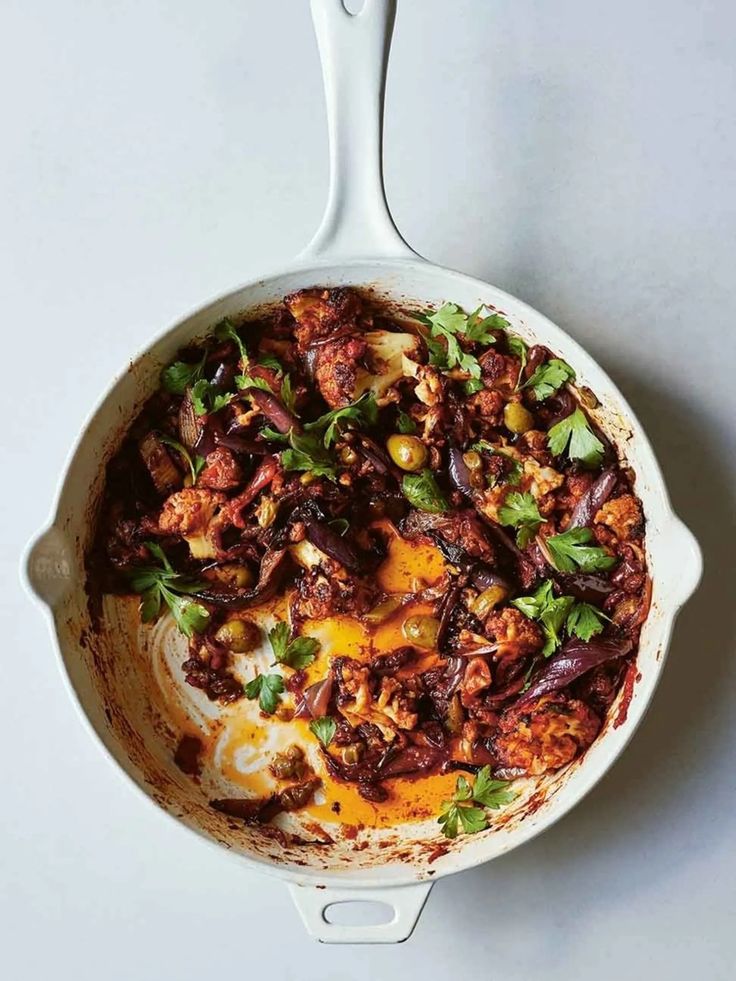 a skillet filled with meat and vegetables on top of a white table next to a spoon