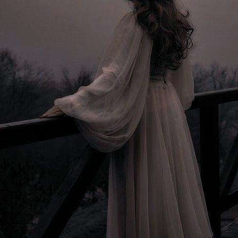 a woman in a long white dress standing on a balcony looking out at the trees