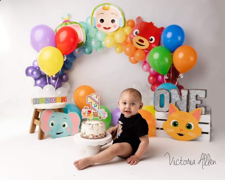 a baby sitting in front of a birthday cake with balloons and animals on the wall