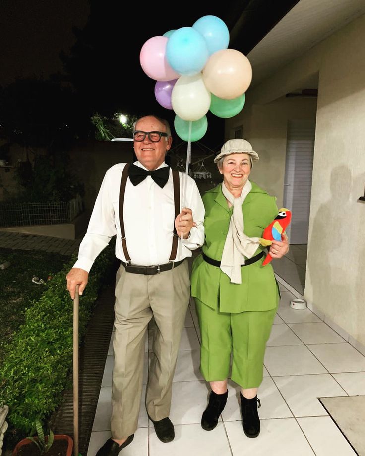 an older man and woman holding balloons in front of a house at night with one person wearing a green outfit