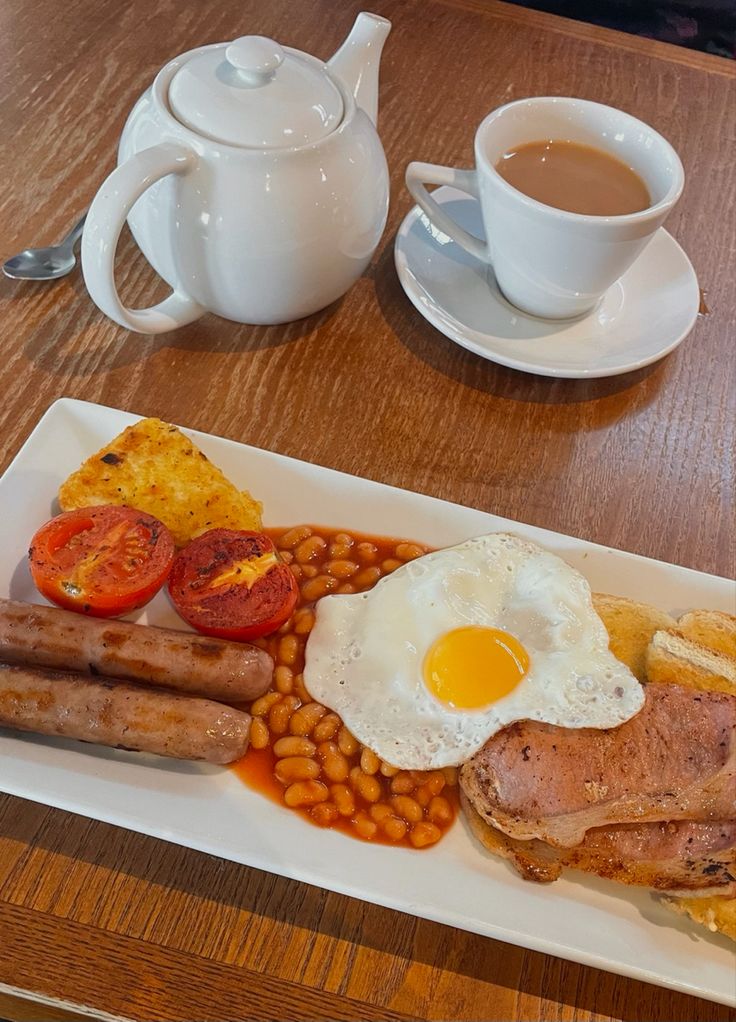 a breakfast plate with eggs, sausages, beans and tomatoes next to a cup of coffee