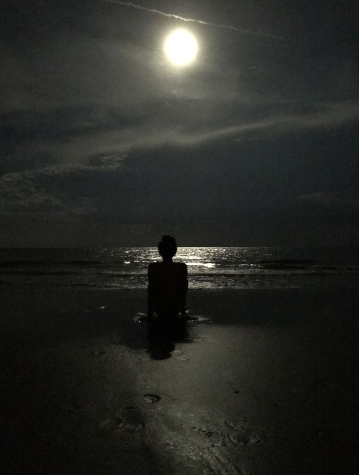 a person sitting on the beach in front of the ocean under a full moon at night