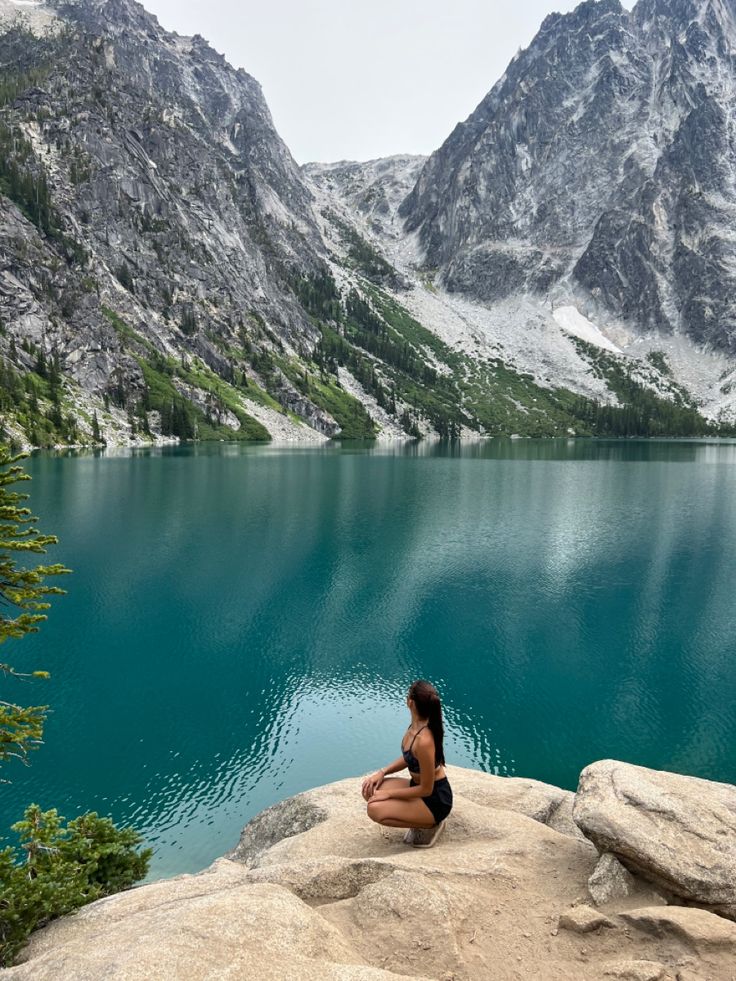 a woman sitting on top of a large rock next to a lake with mountains in the background