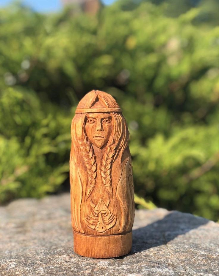 a carved wooden doll sitting on top of a rock next to some bushes and trees