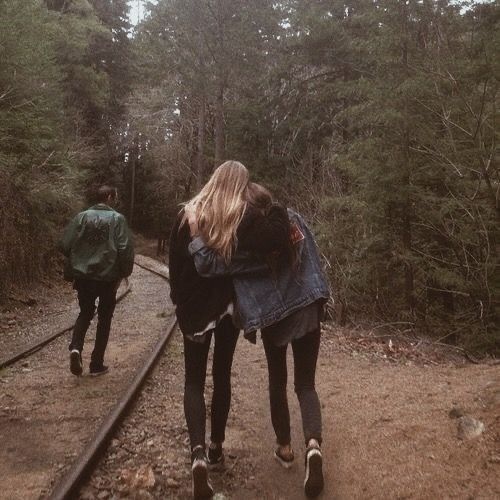 two people are walking down the train tracks in the woods, one is carrying a backpack