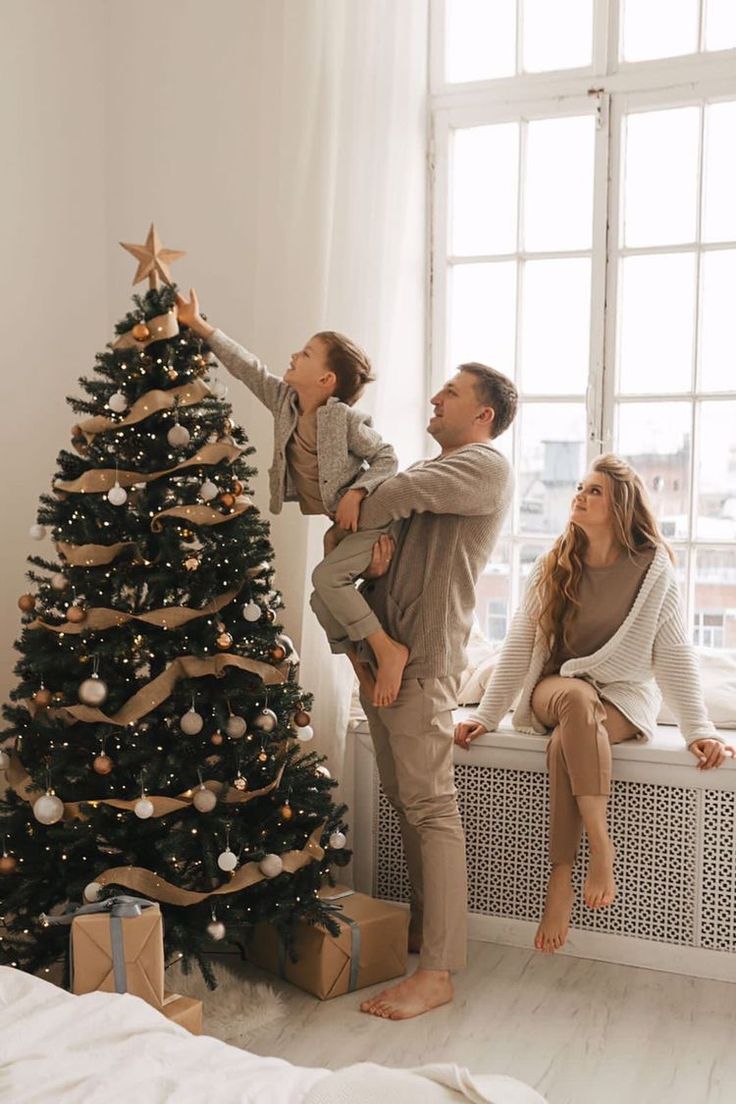 a man and woman are decorating a christmas tree with their daughter, who is sitting on the window sill