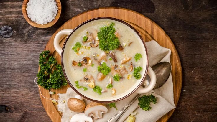 a bowl of soup with mushrooms and broccoli on a wooden plate next to spoons