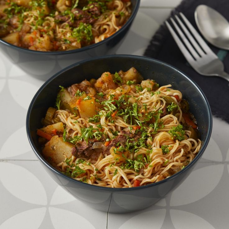 two bowls filled with noodles and vegetables on top of a white tablecloth next to silverware