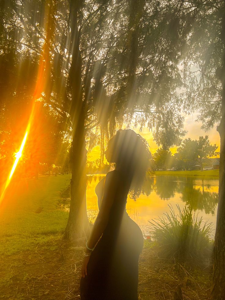 a woman standing in front of trees with the sun shining through her hair and behind her is a body of water