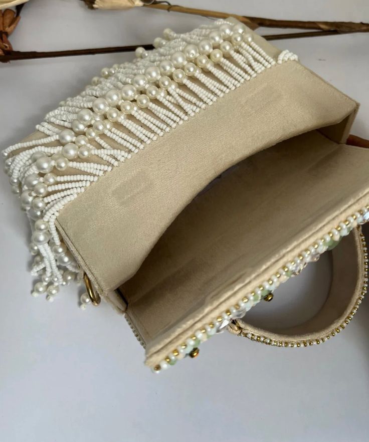 Introducing the Danika Flap Bag, a stunning fusion of bridal elegance and Indian craftsmanship. Hand-beaded with luxurious velvet in enchanting hues of mint, white, and peach, with a beautiful pearl motif, this bag embodies opulence and tradition. Its intricate design, coupled with delicate white suede accents, makes it a standout accessory for brides and lovers of Indian fashion alike. Whether for a wedding ceremony or a cultural celebration, the Danika Flap Bag adds a touch of regal charm to a Cream Pearl Shoulder Bag For Party, Traditional Evening Shoulder Bag With Pearl Handle, Rectangular Beige Bags With Handwork, Beige Rectangular Bags With Handwork, Elegant Hand Embellished Clutch Shoulder Bag, Luxury Embellished White Bags, Luxury White Embellished Bag, Luxury White Embellished Bags, Rectangular Beaded Bag For Reception