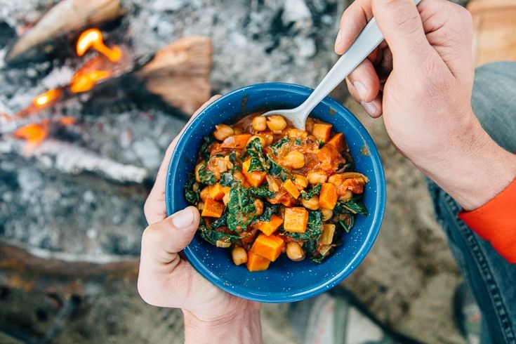 a person holding a blue bowl filled with food next to an open fire and campfire