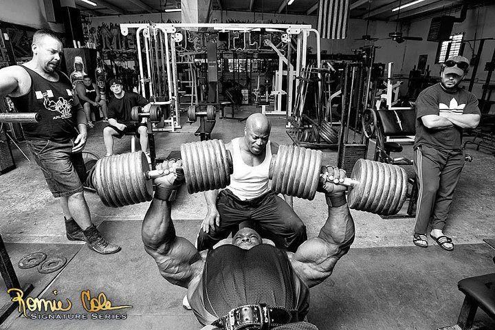 a man doing a bench press with two other men in the background working on their muscles