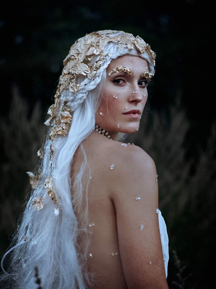 a woman with long white hair and gold jewelry on her face is standing in the grass