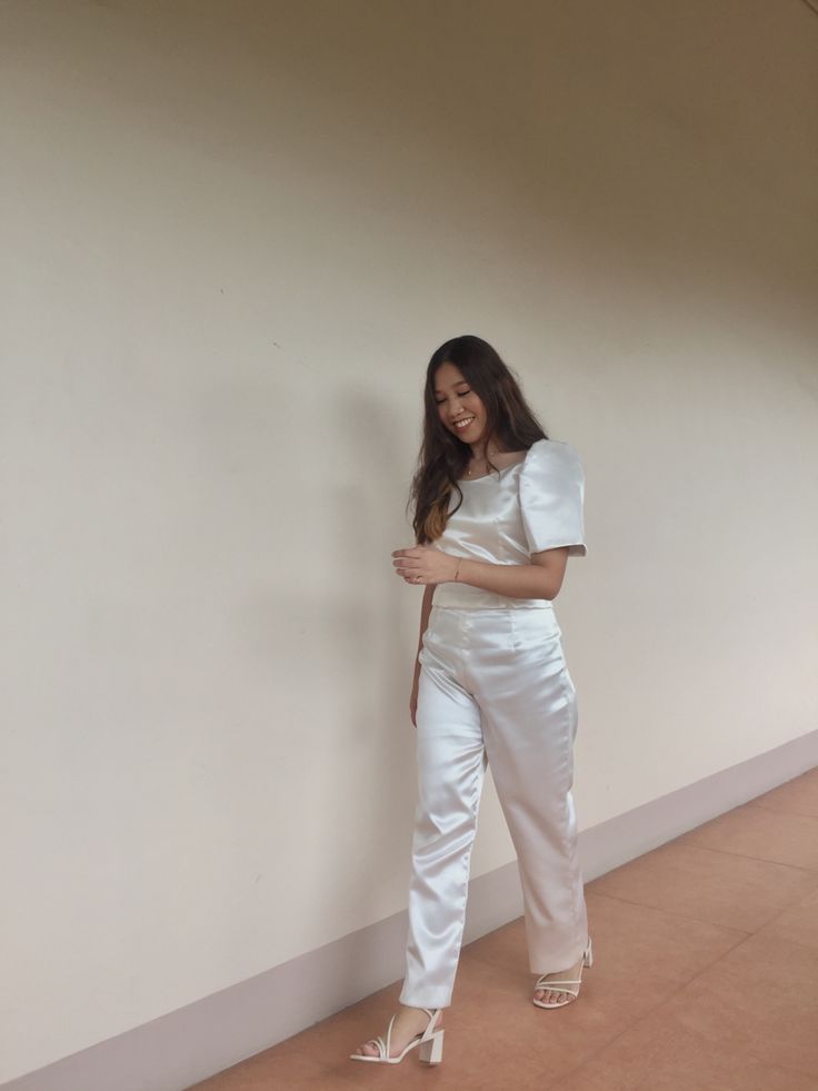 a woman standing next to a wall wearing white pants and a cropped top with one hand on her hip