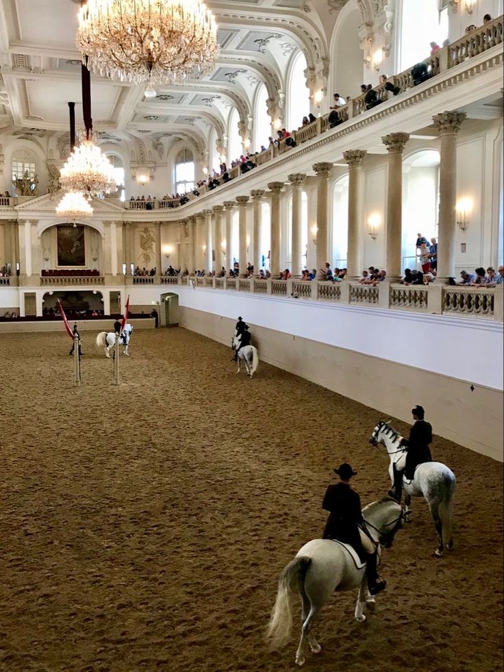 several people riding horses in an indoor arena with chandeliers hanging from the ceiling