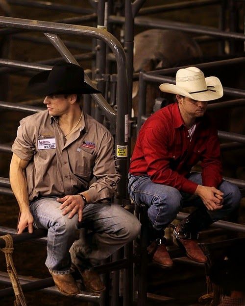 two men in cowboy hats are sitting on some metal bars and one is wearing a red shirt