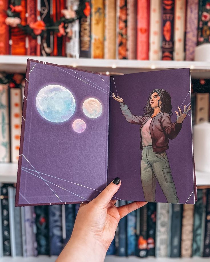 a person holding up an open book in front of bookshelves with moon images on them