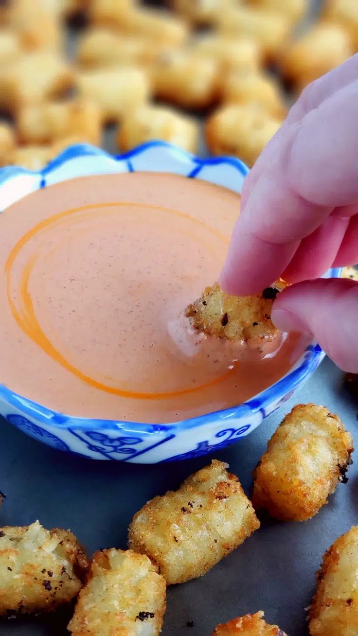 a person dipping something into a bowl of dip with tater tots on the side