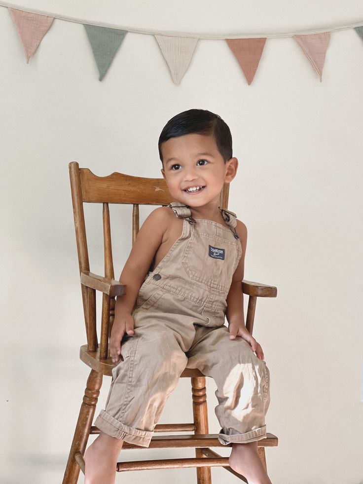 a young boy sitting on top of a wooden chair in front of a bunting flag