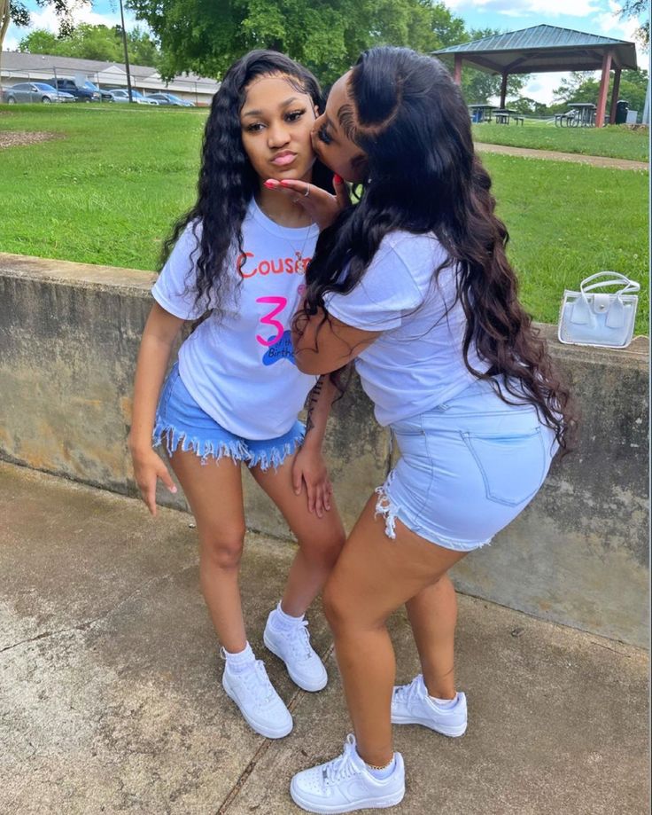 two young women standing next to each other in front of a cement wall and grass